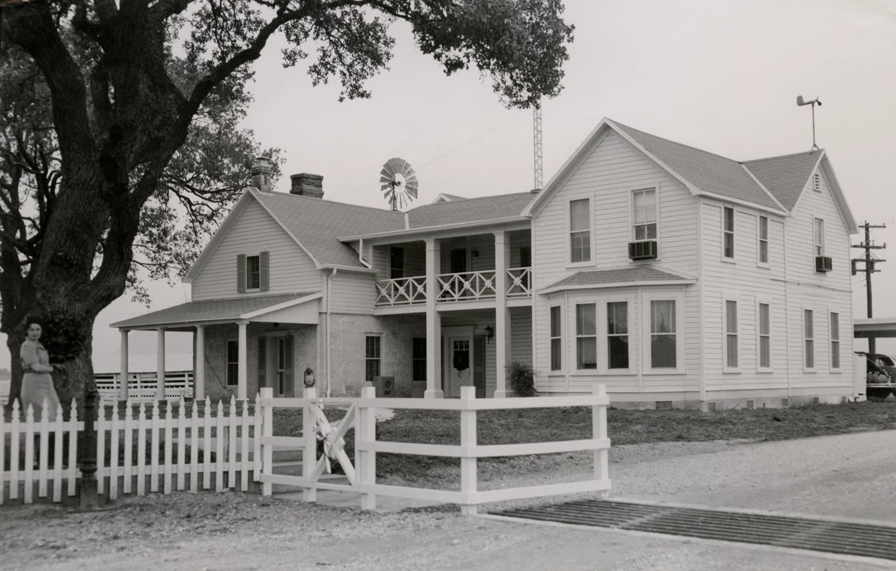 Lady Bird Johnson leans against a tree in the yard of a two-story frame house