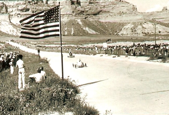 Two soap box cars approach an American Flag as they speed down a road lined with spectators.