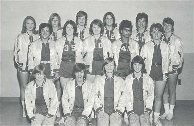 A group of teenage girls in basketball uniforms. One girl is African-American and all the others are white.