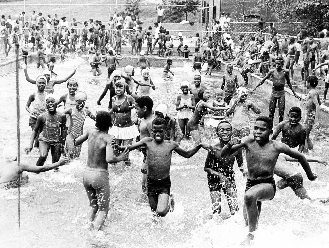 A crowd of Black kids joyfully splash in a large outdoor pool as others wait along the edge.