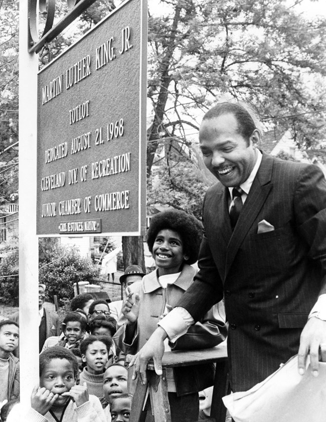 Dressed in a suit, Stokes stands on a stepladder beside a park sign, Children smile below him.