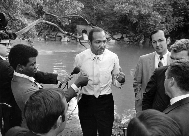 African American man is surrounded by reporters along a river.