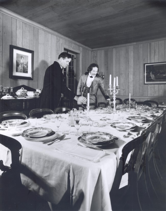 1970s candlelight A black and white photo of two re-enactors setting the mess hall dining table inside the Chief Factor's House at Fort Vancouver.