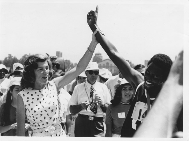 Eunice Kennedy Shriver stands with a black athlete. They have their hands raised, Eunice holding the athlete's wrist.