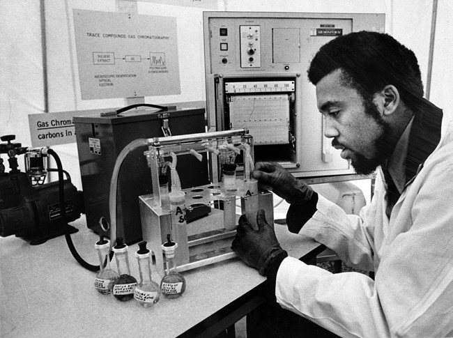 A Black man in a lab coat leans over test tubes and vials. The rack attaches to recording equipment.