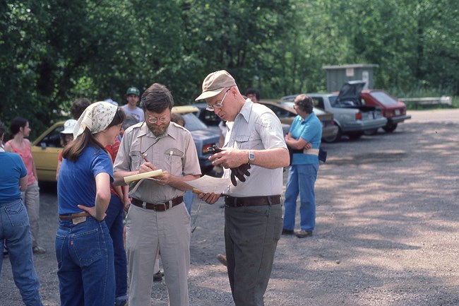 Volunteers in work clothes stand chatting in a wooded lot by a line of parked cars.