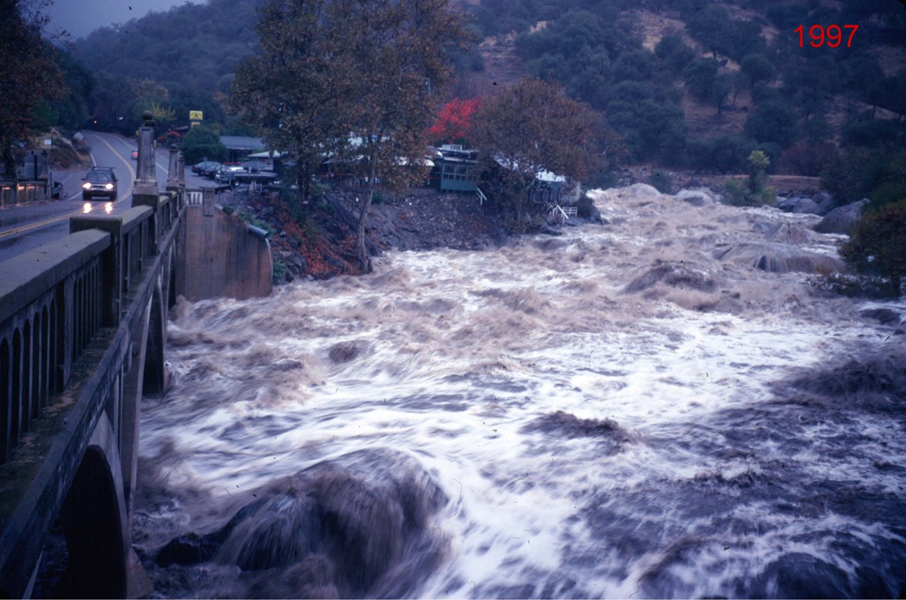 Late afternoon view from bridge of muddy, high river flowing so high that it covers most of the large boulders that are usually exposed.