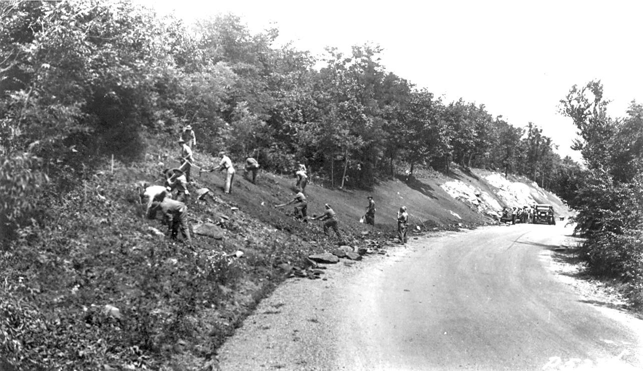 Black and white photo, a group of workers with tools dig on a cleared slope to the left of a roadbed