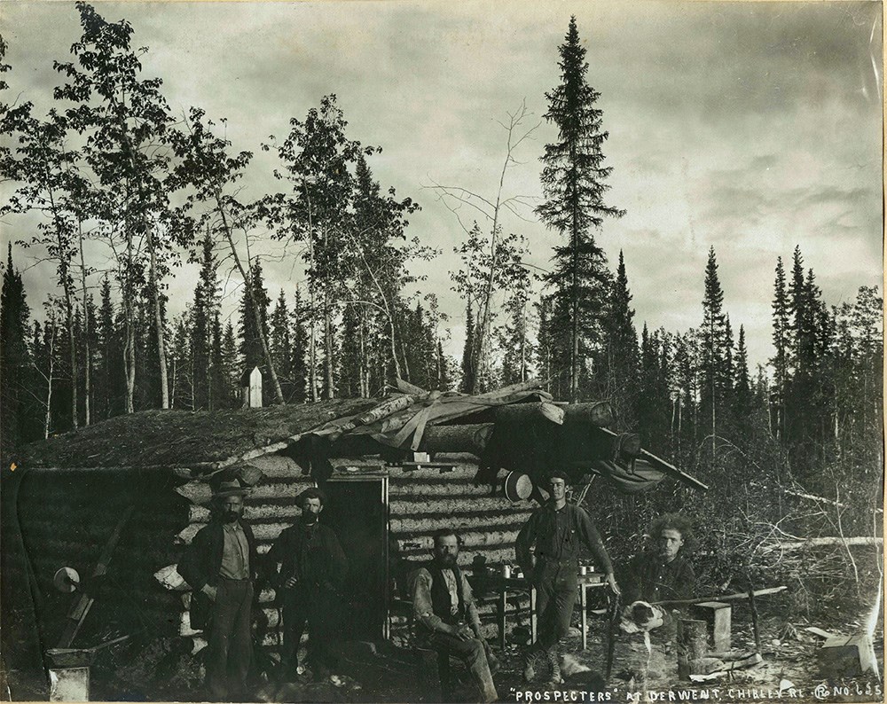 Historical black and white photo of prospectors by their log cabin.