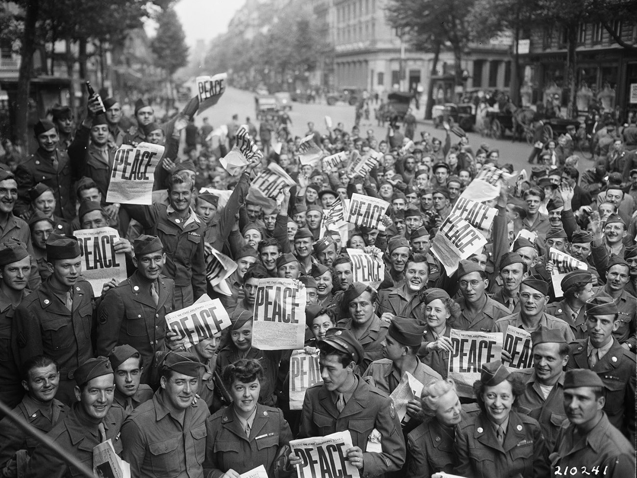 lt= Many soldiers in uniform crowd a street holding newspapers that announce "Peace."