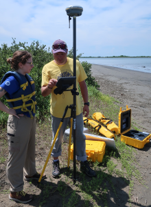 Two people staring at the screen display of a drone while the drone is operating on the beach of the marsh island.