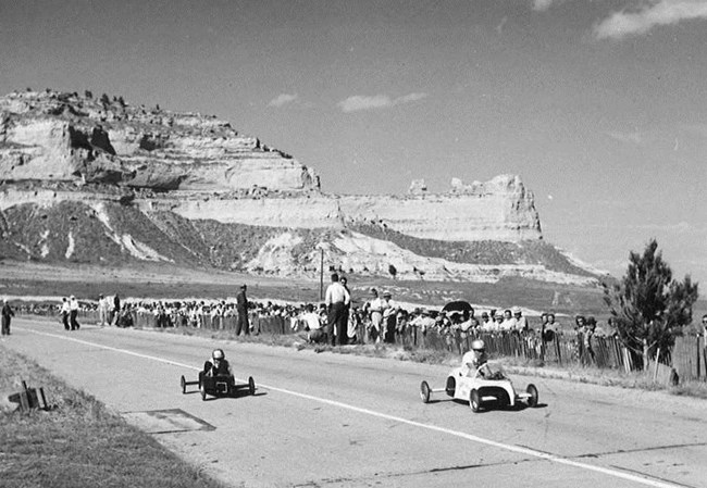 Two soap box cars race down a hill with a crowd of spectators and sandstone bluff in the background.