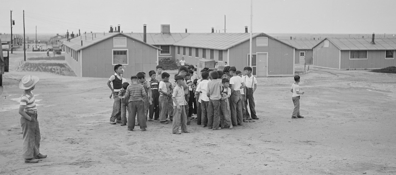 Children playing at Amache A group of young boys congregate on a dirt field in front of a large complex of buildings.