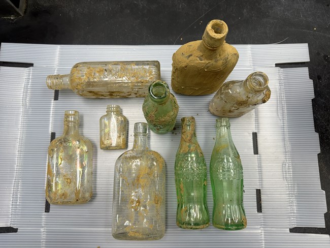 Nine old glass bottles covered in mud on a display table.