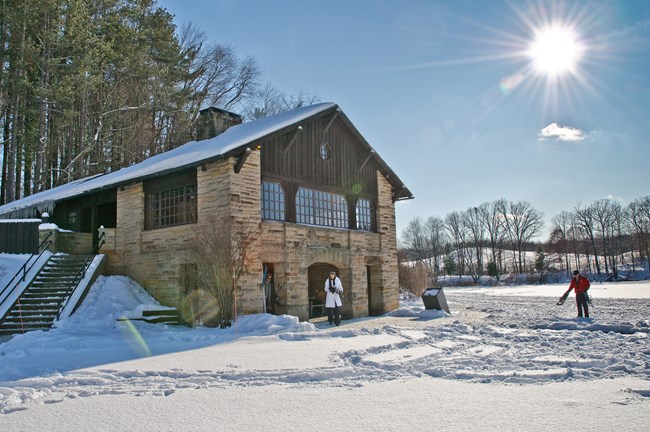 Exterior of a two-story building of gray stone and dark brown wood, on a sunny day with snow on the ground.