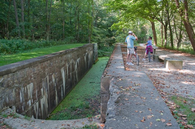 Two people stand with bicycles by a bench next to a canal lock; the lock is two parallel gray, concrete walls; green water sits at the bottom of the lock.