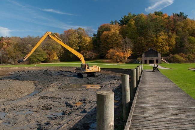 A photo of a yellow excavator down in the muddy lake bed; a wooden boardwalk on the right leads back to the stone shelter.