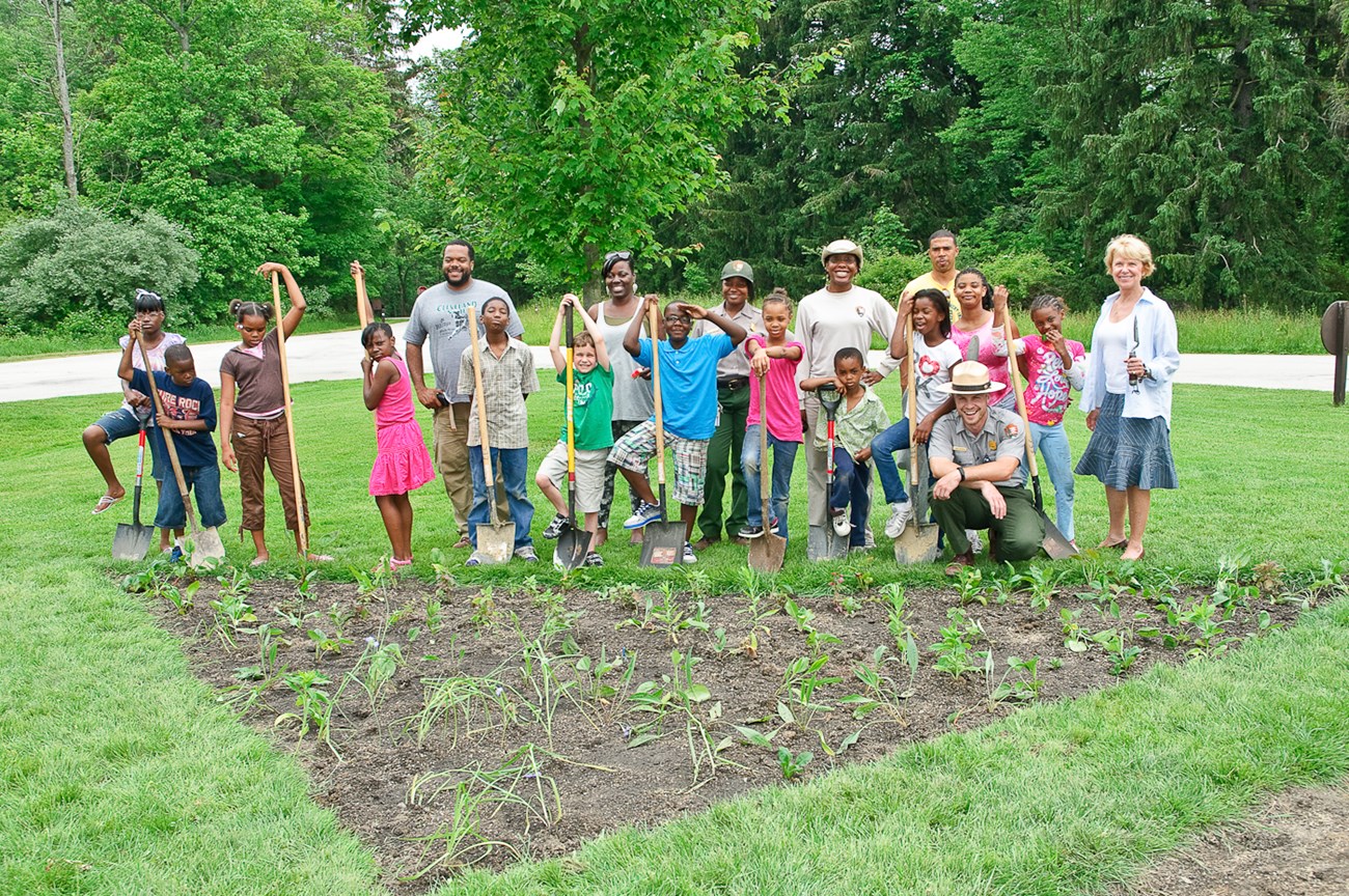 A group of about twenty mostly Black children and adults stand in a line smiling and holding shovels; they stand in front of a triangular patch of freshly planted seedlings.