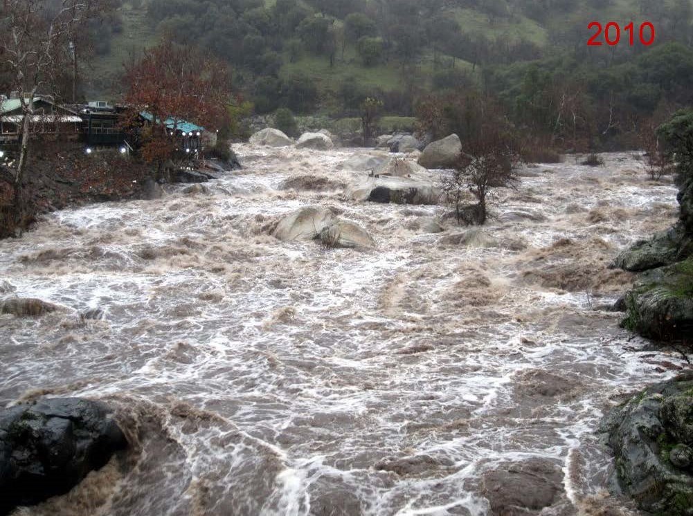 View from bridge of murky river water rushing over most large boulders, and flowing  at high level, bank to bank in river channel.