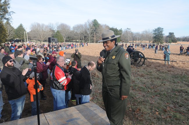 A Black female ranger in formal green uniform holds a microphone and smiles; behind her, hundreds of people stand along the edge of a field full of costumed reenactors and cannons.