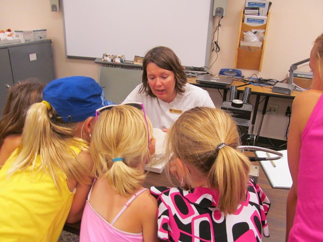 children gather around a woman who sits at a table with a microscope.