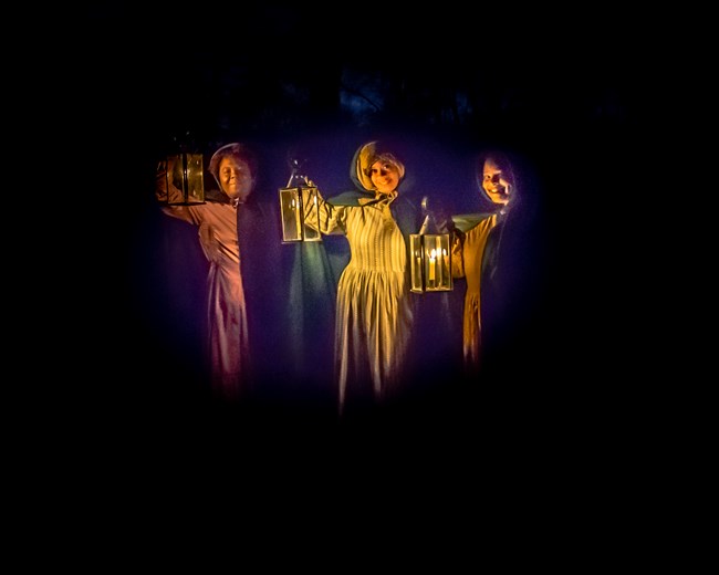 Three Black women dressed in 1800s costume stand in the dark holding candle-lit lanterns.