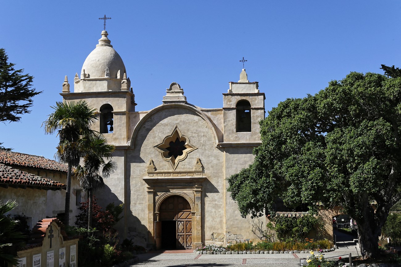 Sand colored stone church has central wooden door, flanked by two towers, topped with wrought iron crosses. Sits in enclosed Spanish-style courtyard