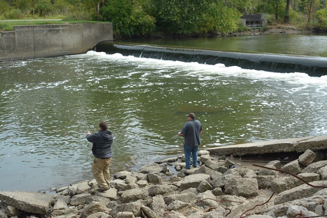 20151006 CRWT-SRB dam fishing Two people fish from the near, rocky shore of the river; upstream of them water flows over the dam.