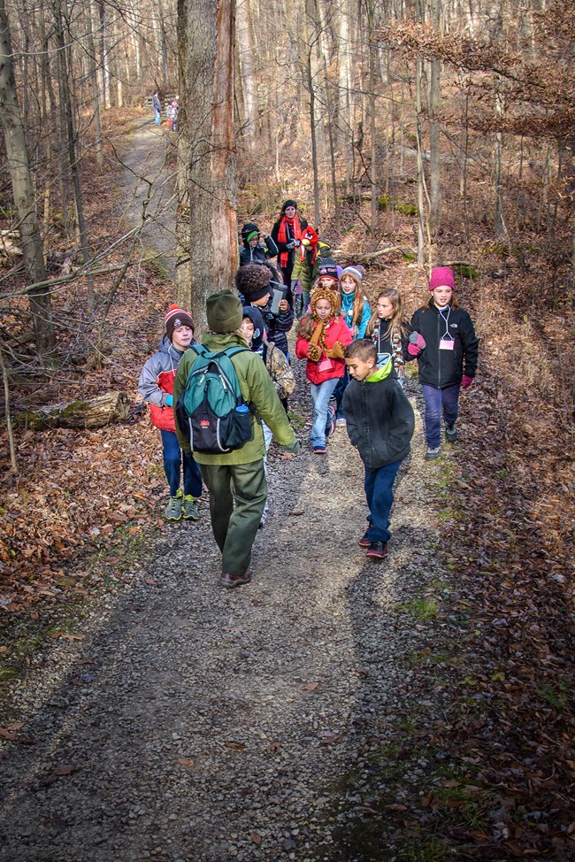 A uniformed ranger in green jacket and hat walks backward down a wooded trail in winter; a dozen children in winter coats and hats follow her.