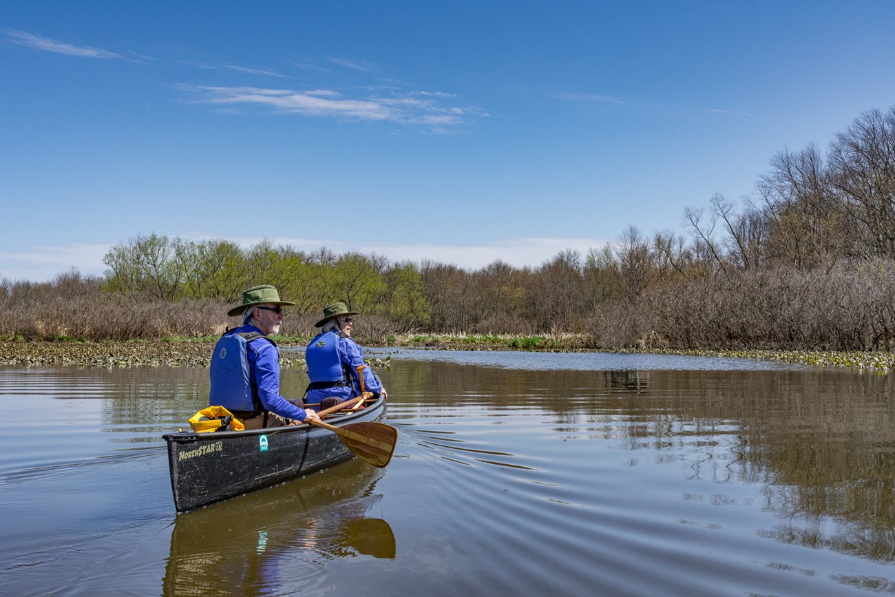 A couple with white hair, green hats, and blue life vests paddle a canoe on a still pond in spring.