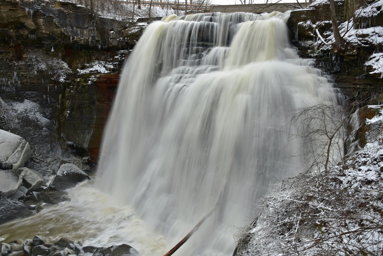 Long-exposure photo of yellow-white water falling over a gray rock face; snow and ice coat the rocks and shrubs on either side.
