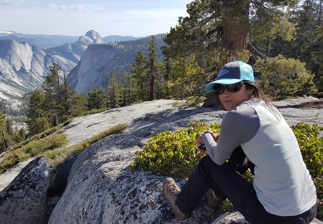 Woman sits on granite rock with view of Yosemite Valley, looking back over her shoulder at the camera.
