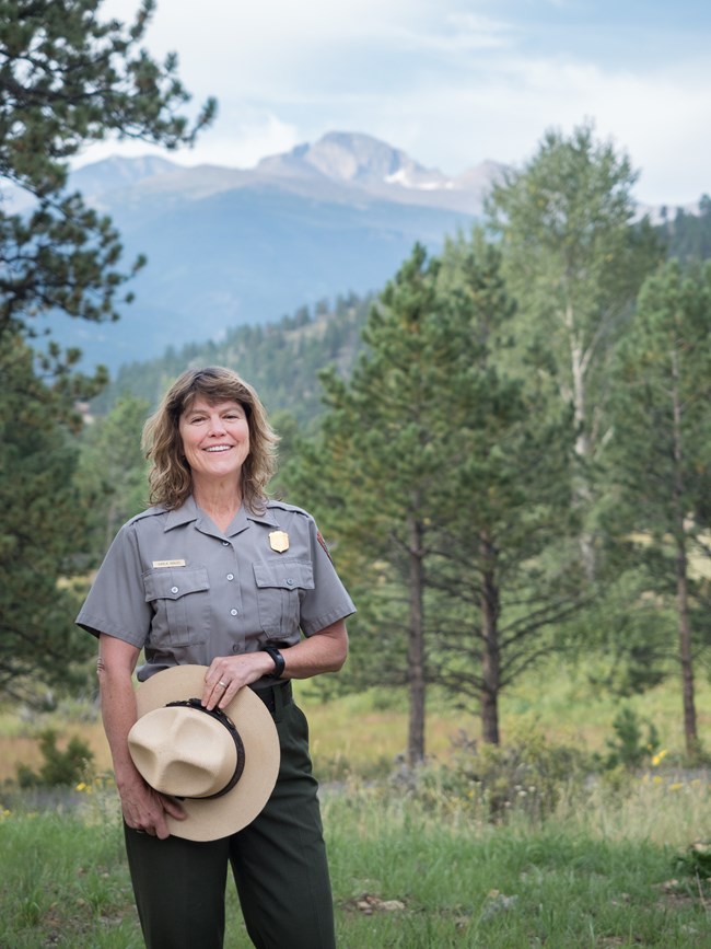 a woman stands in front of a mountain view holding a flat hat