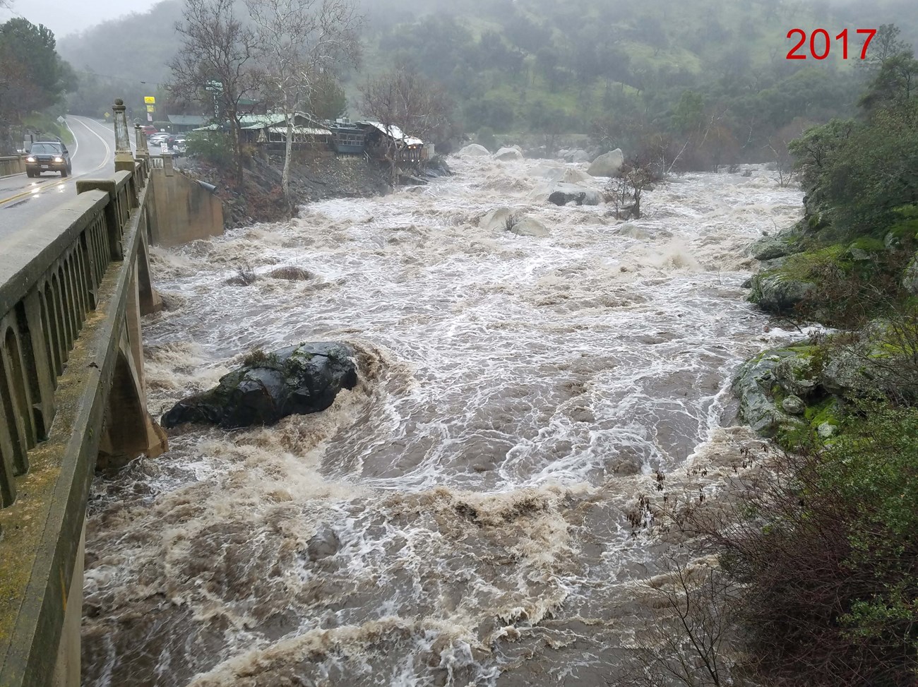 View from bridge of bank to bank muddy river water covering most big rocks.