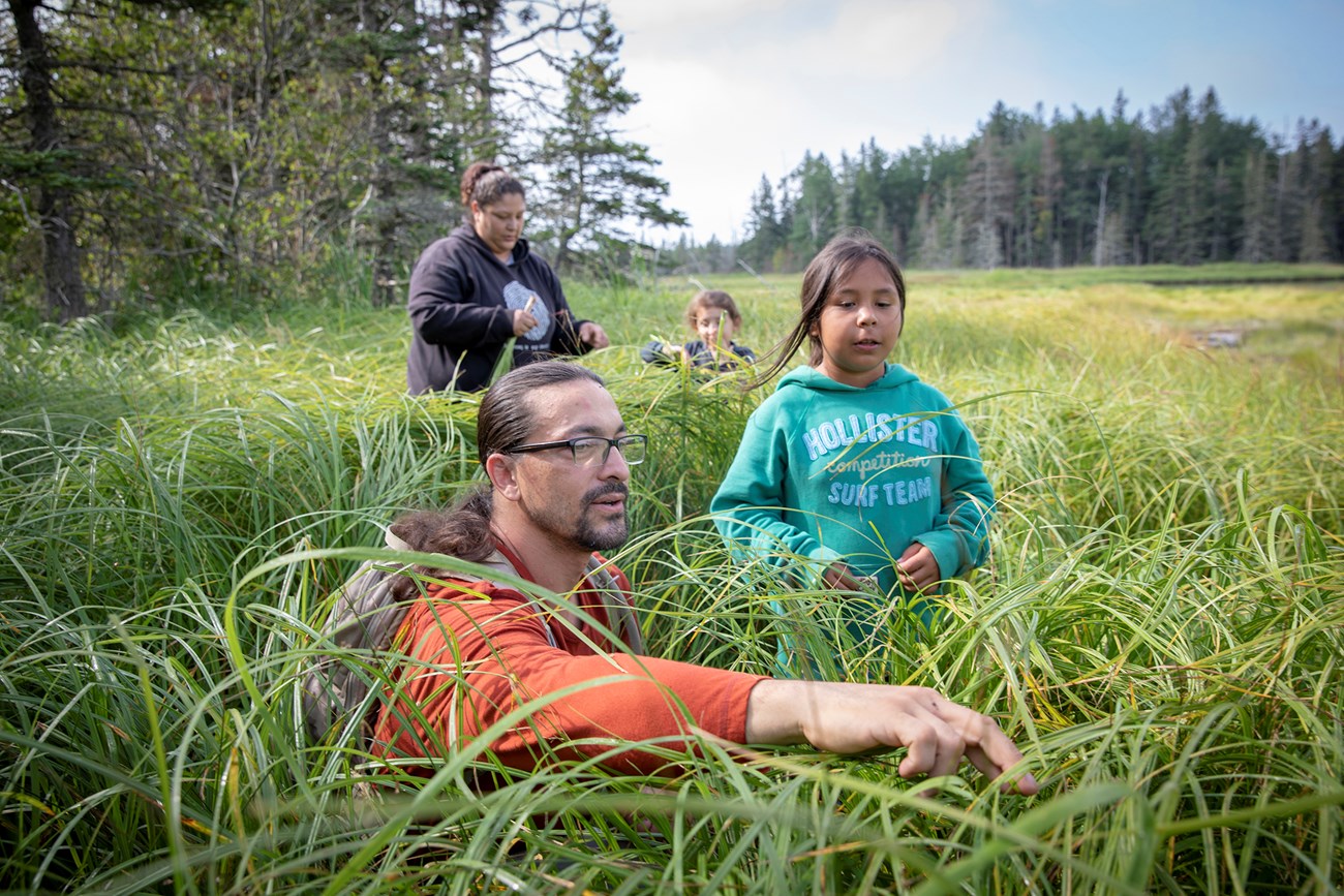 Family standing in a field of grass, one man in the foreground points to a snail. A child is next to him. A woman stands in the background. Trees are in the distance. It's a sunny day.