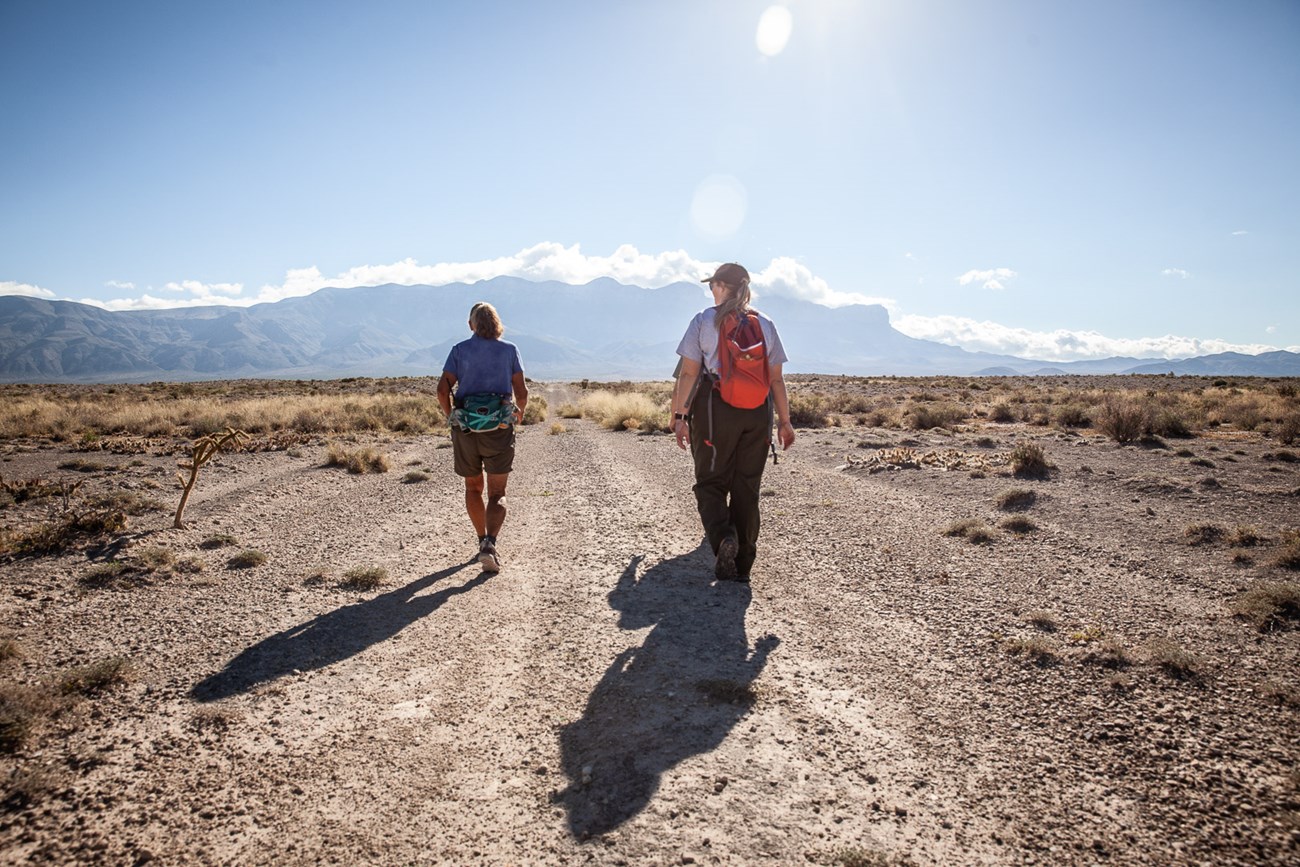A woman and a park ranger walk on a flat trail towards tall mountains A woman and a park ranger walk on a flat trail towards tall mountains