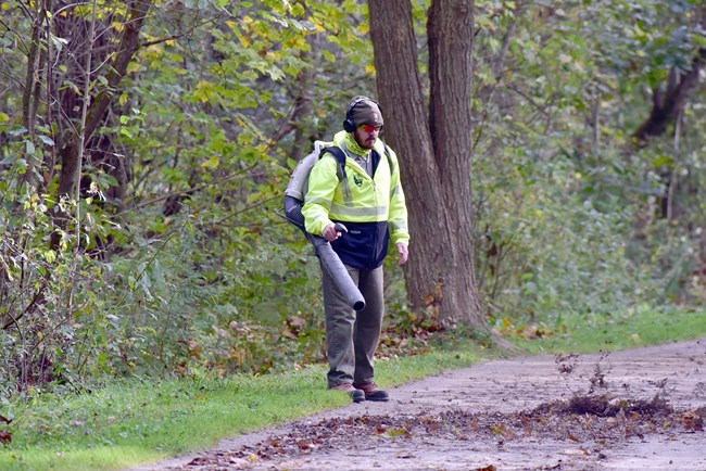 Uniformed ranger wearing a bright yellow sweatshirt holds the nozzle of a backpack blower, blowing leaves from a wide, gray trail with trees in the background.