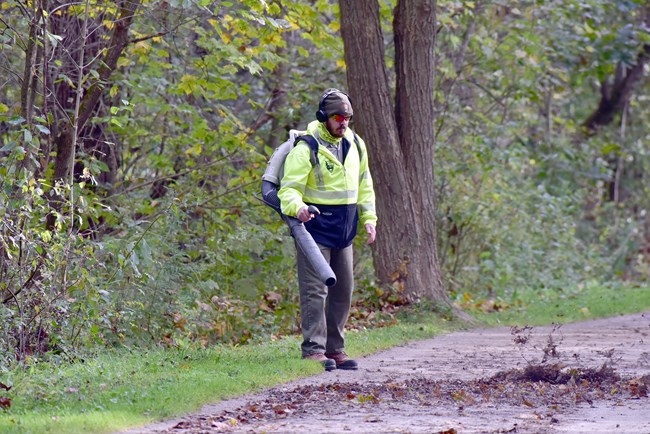 20181019 TT-BM trail maintenance Uniformed ranger wearing a bright yellow sweatshirt holds the nozzle of a backpack blower, blowing leaves from a wide, gray trail with trees in the background.