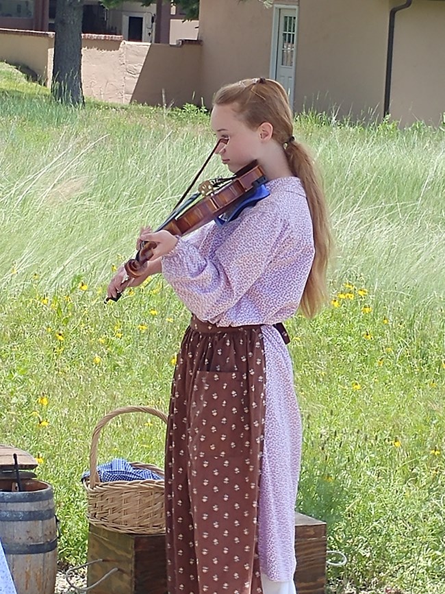 A young lady wearing a sun dress and bonnet plays a violin.