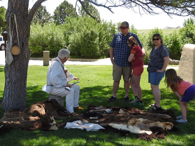 A man dressed as a fur trader talks to a family of visitors in the shade of a tree.