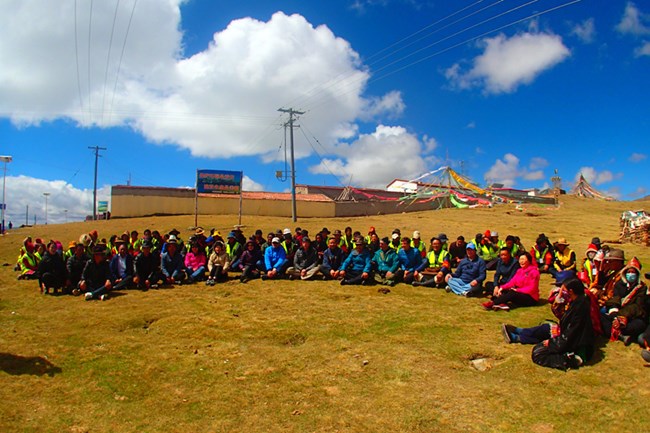 a large group of people sit on a grassy rise with buildings in the background and puffy white clouds in a bright blue sky