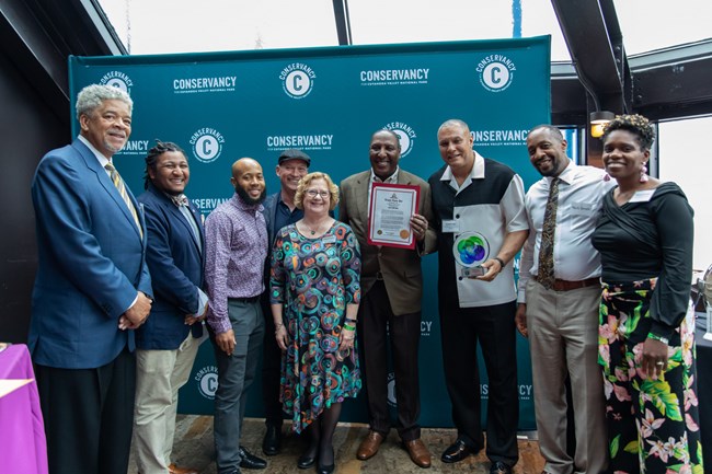 Nine people pose in front of a Conservancy banner. One man holds an award statue and another a certificate.