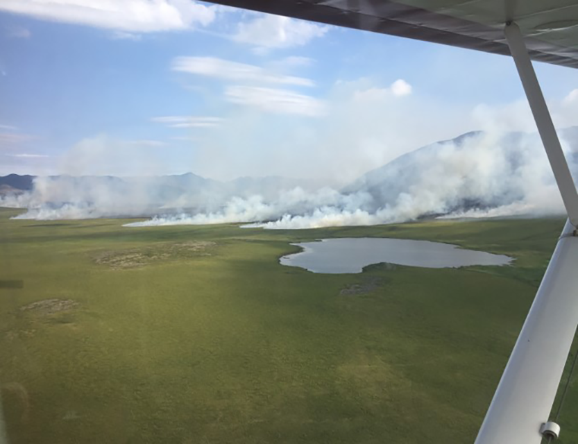 An image from an aircraft of the 2019 Kaluktavik River Fire burning in a tundra landscape with ringed by mountains and with a small lake in the foreground.