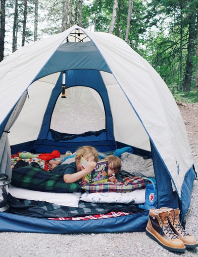 Two children read a book inside a tent pitched in a park campground.