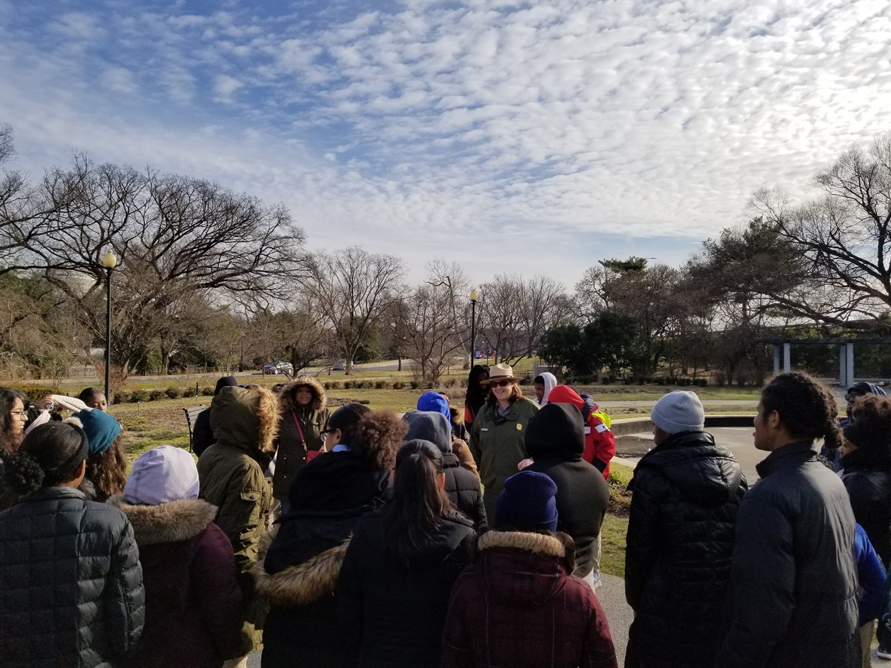 A group of students stand in front of a park ranger in uniform at the George Mason Memorial with blue sky and clouds above.