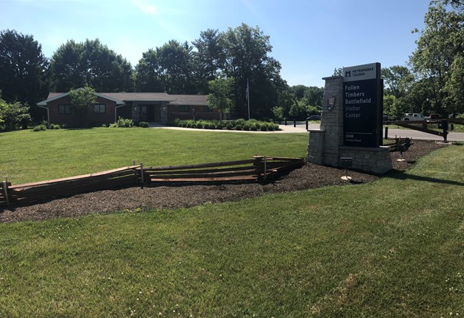 A building in the back with a grassy yard. In the forefront is a welcome sign that reads "Metroparks Toldedo: Fallen Timbers Battlefield Visitor Center" with the National Park Service emblem to the left on the sign's pillar.