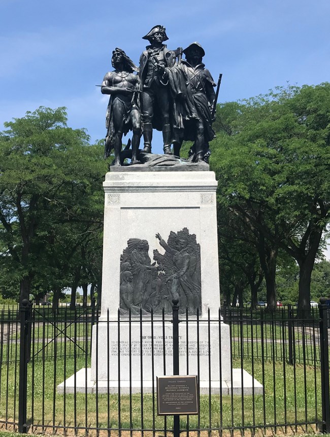 A bronze statue with three figures sits on a tall stone base with a Greenville Treaty relief.