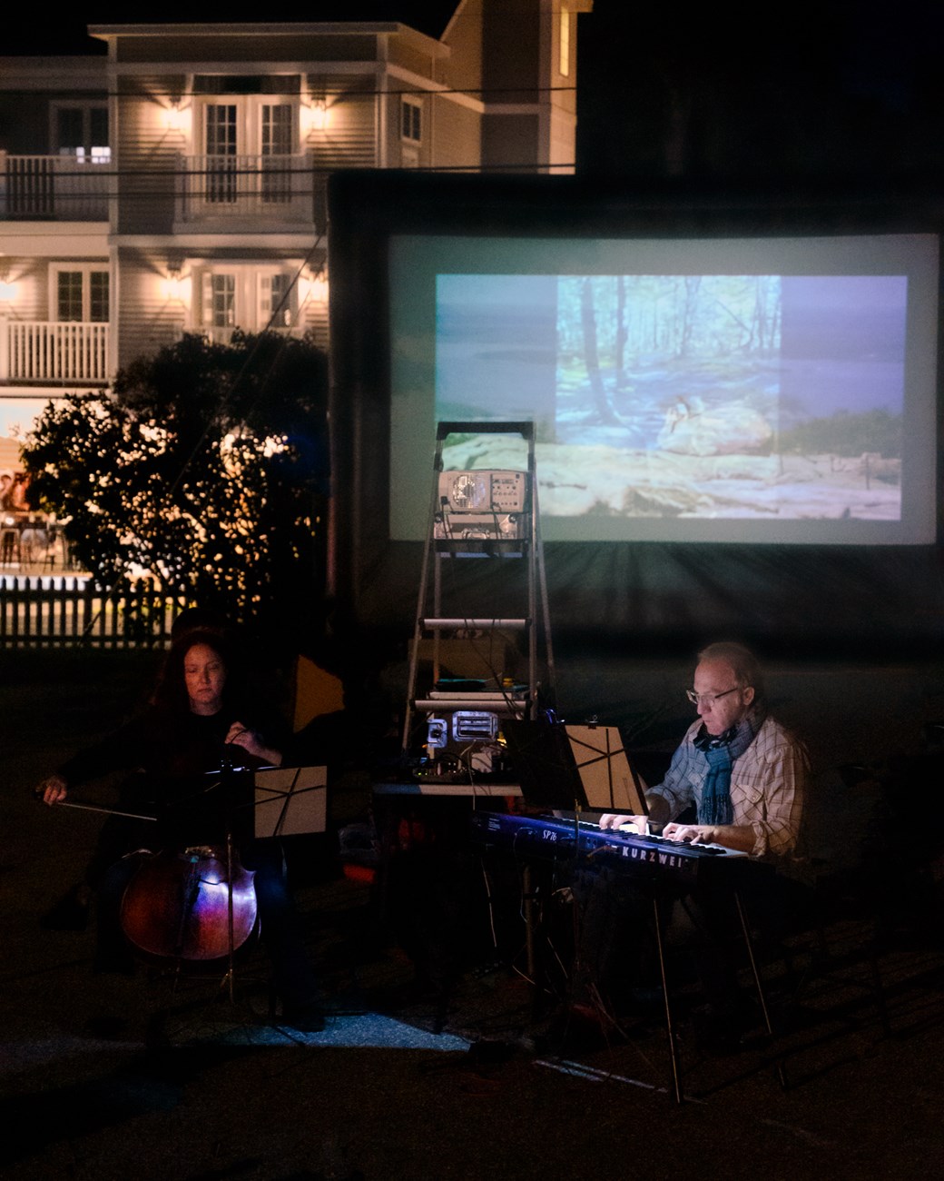 In a dark outdoor space in front of a movie screen, a man plays piano and a woman plays cello.