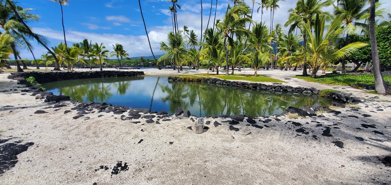 A small fish pond with stacked lava rock walls sits amid the white coral sands and coconut trees of the Royal Grounds.