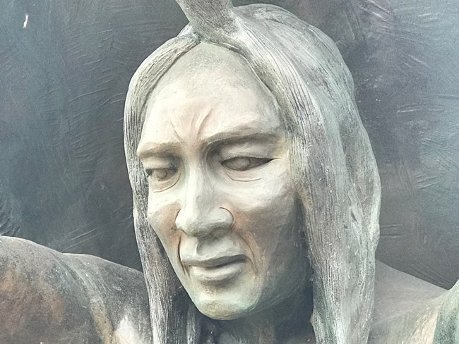 Face of a man with long hair and a topknot carved in bronze.